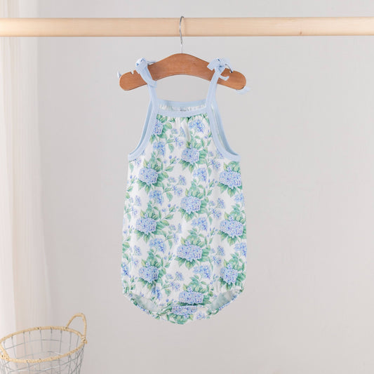 Nola Tawk Hydrangea Heaven baby onesie featuring a blue and green floral print with adjustable shoulder ties on a wooden hanger.