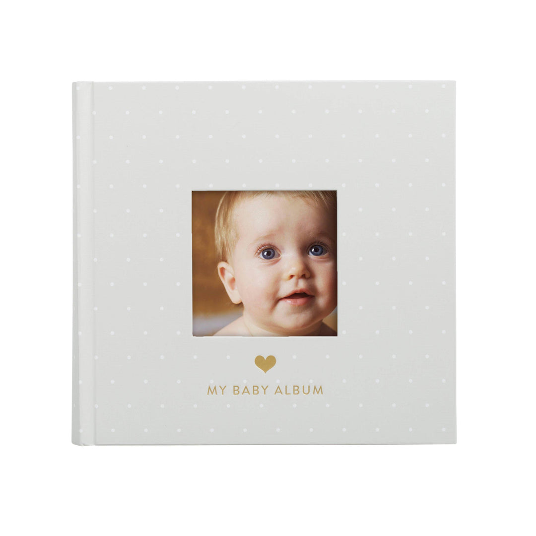 A baby photo album with a white cover decorated with small gold polka dots, featuring a central square cutout showing a baby&