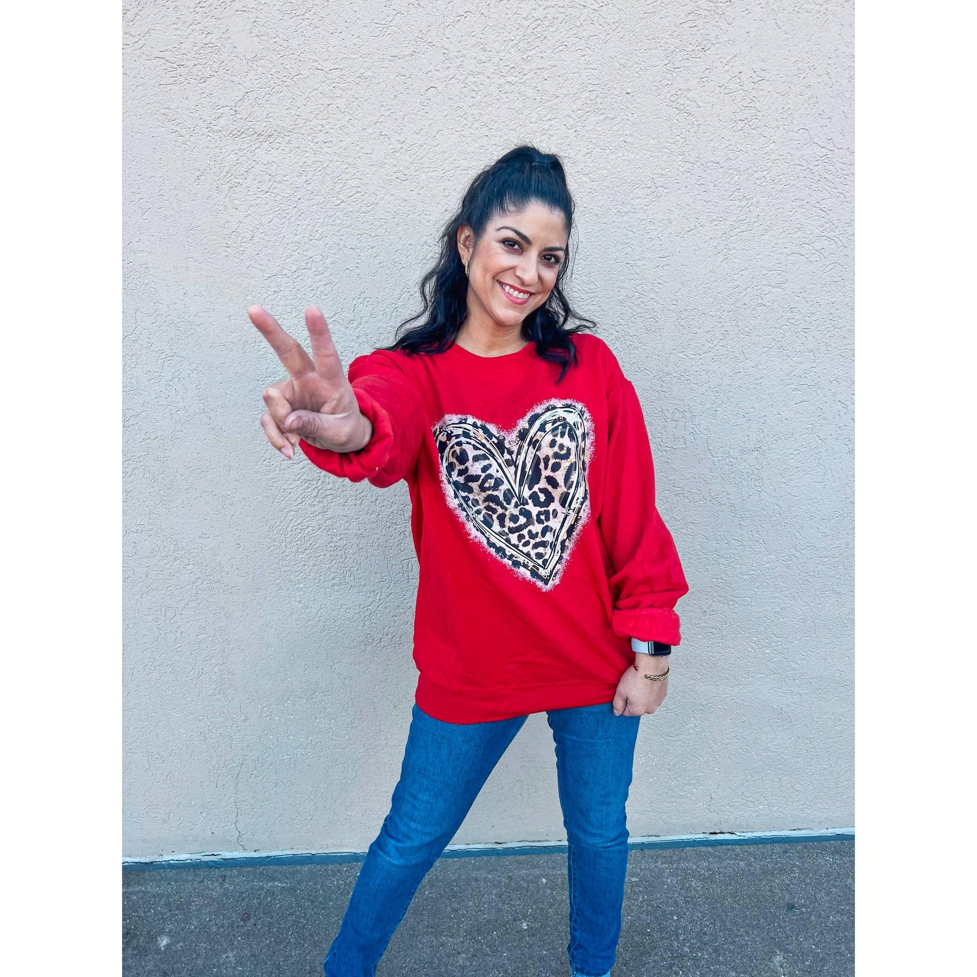 Woman wearing a red sweatshirt with a heart-shaped leopard print design, standing against a light gray wall.