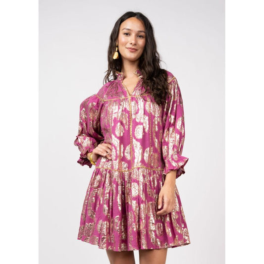 A woman wearing a magenta swing dress with gold metallic rose detailing, long ruffled sleeves, a split neckline, and a tiered skirt.