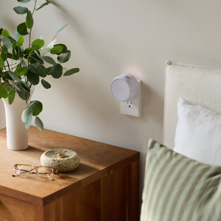Pura Home diffuser plugged in beside a bed and nightstand in a bedroom