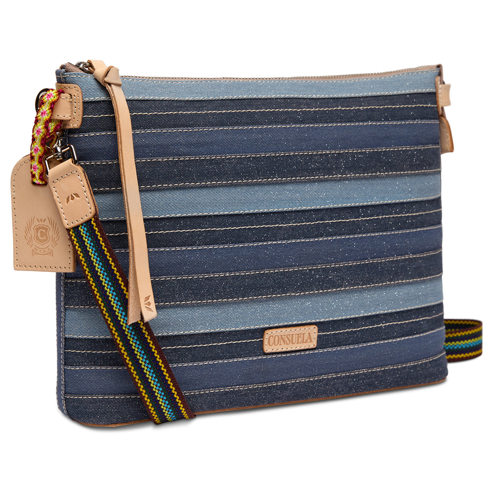 Three-quarter angled view of the Consuela Royal Downtown Crossbody showing the blue denim-stripe fabric, side depth, and colorful woven strap.