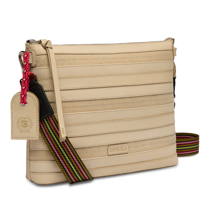 Three-quarter angled view of the Consuela Amina Downtown Crossbody in natural leather with horizontal stitched panels and a colorful rainbow woven strap.
