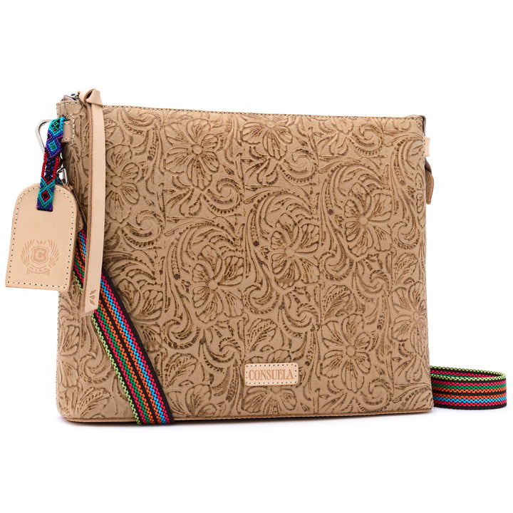Consuela Lawton Downtown Crossbody in neutral tan tooled floral leather, three-quarter front view with a colorful woven strap and leather hangtag.
