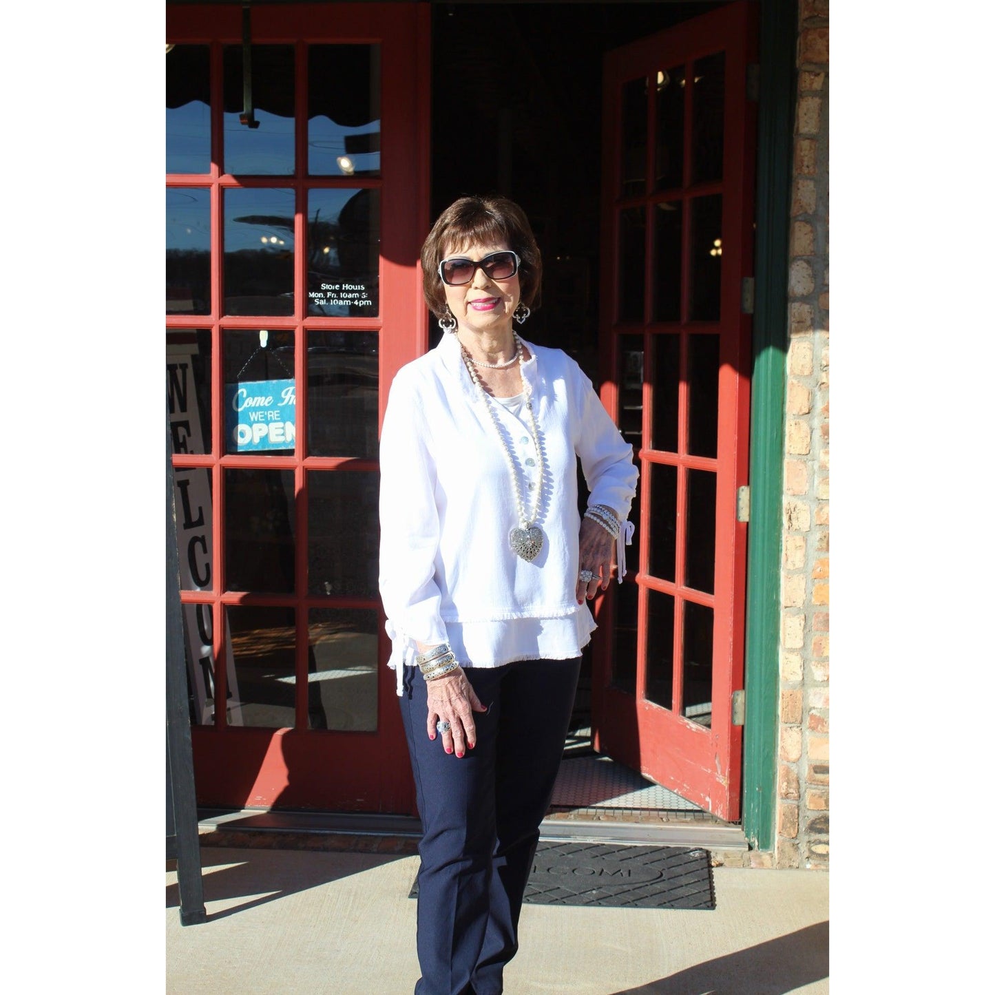 Woman standing in front of a red door with glass panels, wearing a white coat and sunglasses.