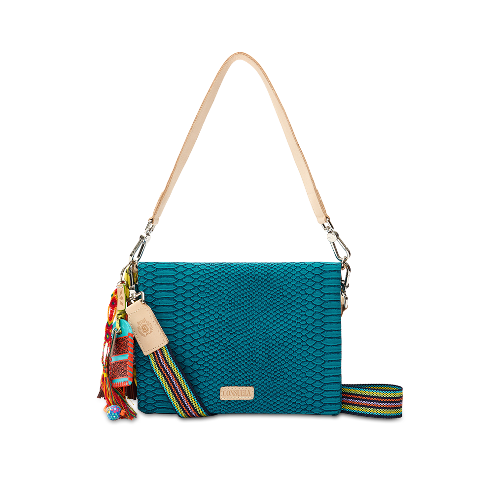 Consuela Indy Metro Crossbody in teal reptile-embossed leather, front view with a charm tassel and multicolor woven crossbody strap and a leather shoulder strap on a checkered background.