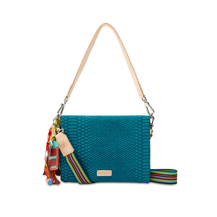 Consuela Indy Metro Crossbody in teal reptile-embossed leather, front view with a charm tassel and multicolor woven crossbody strap and a leather shoulder strap on a checkered background.