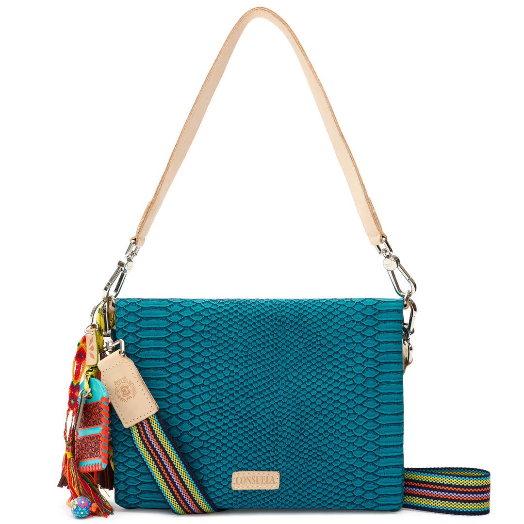 Consuela Indy Metro Crossbody in teal reptile-embossed leather, front view with the flap closed, a natural leather strap, a colorful charm tassel, and a woven crossbody strap.