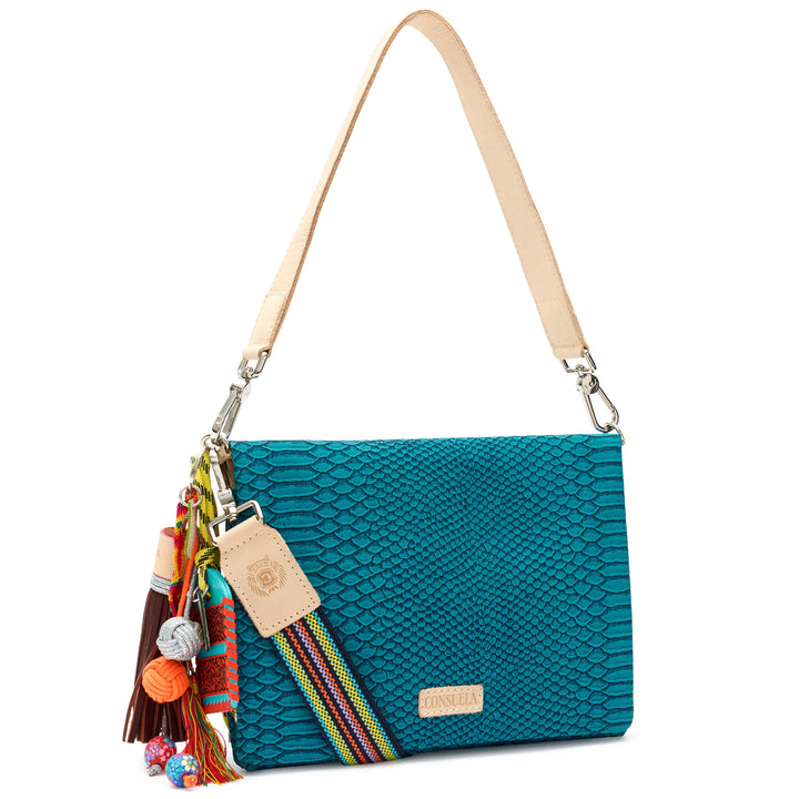 Three-quarter angled view of the Consuela Indy Metro Crossbody in teal reptile-embossed leather with silver hardware and a colorful charm tassel.