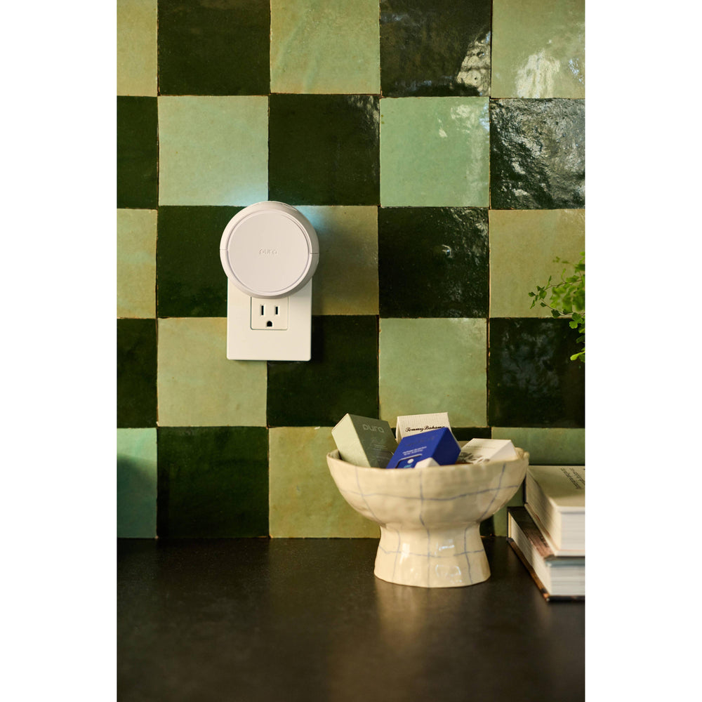 Pura Home smart diffuser plugged into a green checkered tile wall in an entryway