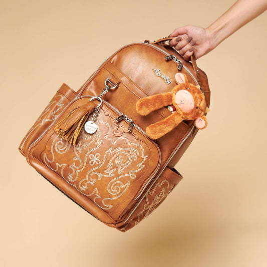 "Itzy Ritzy Boss Plus™ diaper bag backpack in Nash Saddle brown vegan leather with western-style stitching and a plush lion bag charm accessory."

