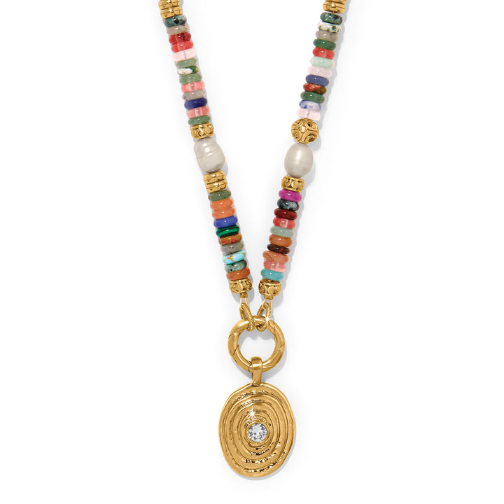 The Brighton Kosmos Chroma Token Charm Necklace displayed on a white background. It features a chain made of colorful semi-precious style beads and faux pearls, leading to a textured, gold-tone oval pendant with a clear crystal in the center.